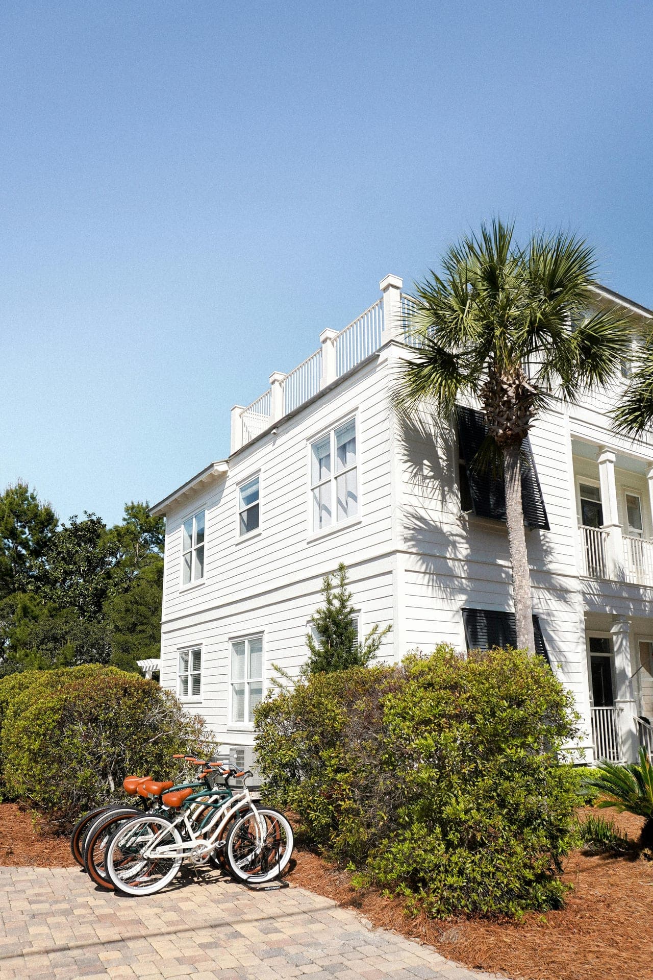 A white 30A beach house framed by a palm, with beach-cruiser bikes parked out front — the kind of Emerald Coast rental owners enroll.