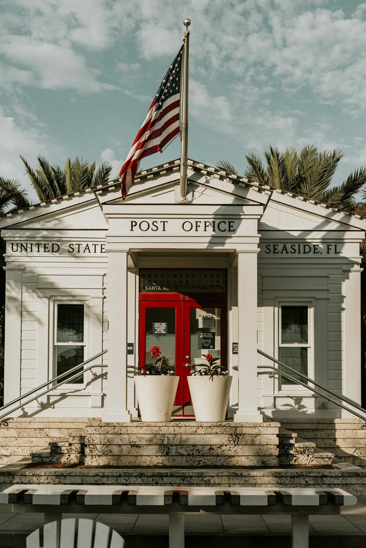 The historic Seaside, Florida Post Office — American flag, white pediment with POST OFFICE / UNITED STATE / SEASIDE, FL. signage, red door, and planters at the steps.